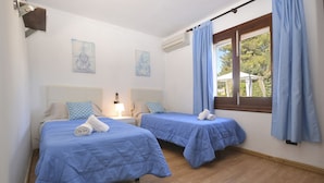 A room with two beds, each with a blue bedspread and a rolled towel. There are two wall-mounted air conditioning units, a window with blue curtains, and two wall-mounted paintings. - Llimona - Cozy house with pool in Alcúdia, Mallorca (Alcúdia)