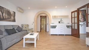 A modern living room with a grey sofa, a white coffee table, and a kitchen area with white cabinets and a wooden door. - Llimona - Cozy house with pool in Alcúdia, Mallorca (Alcúdia)