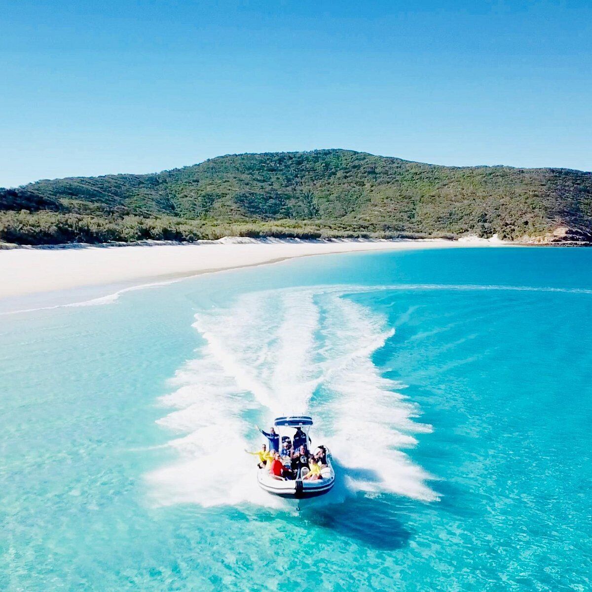 A boat with passengers on a clear turquoise sea, surrounded by a sandy beach and a green hill in the background.