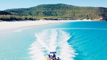 A boat with passengers on a clear turquoise sea, surrounded by a sandy beach and a green hill in the background.
