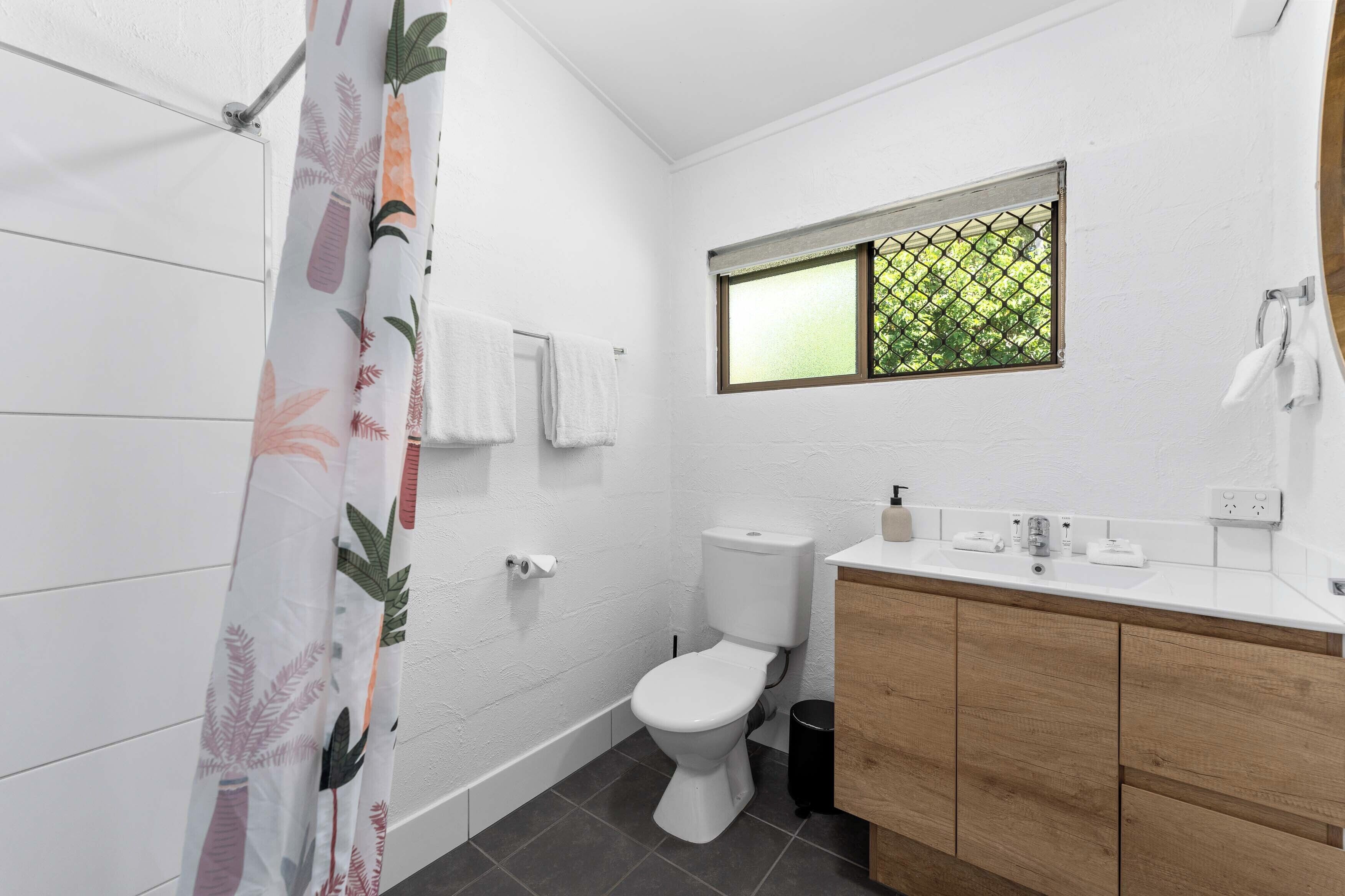 A modern bathroom with a white tiled wall, a wooden vanity, a toilet, a window with a view, and a shower curtain with a leaf pattern.