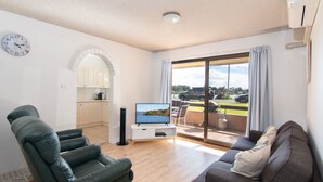 Un salon moderne avec un téléviseur à écran plat, un canapé et un fauteuil. On y trouve également un coin cuisine avec des meubles blancs et une horloge au mur.