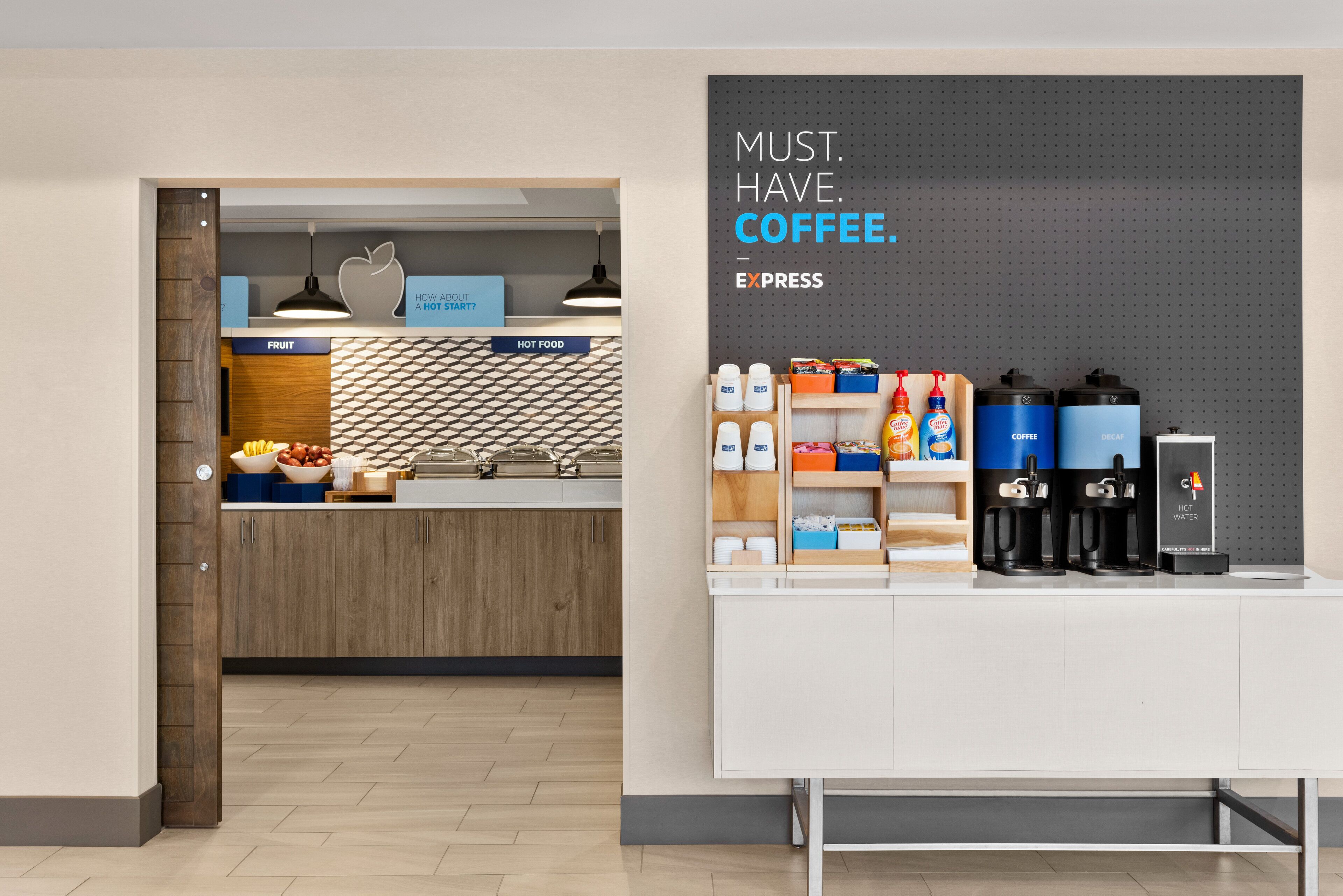 A modern coffee station with a sign reading "MUST HAVE COFFEE - EXPRESS" and a wall-mounted pegboard with various coffee products.