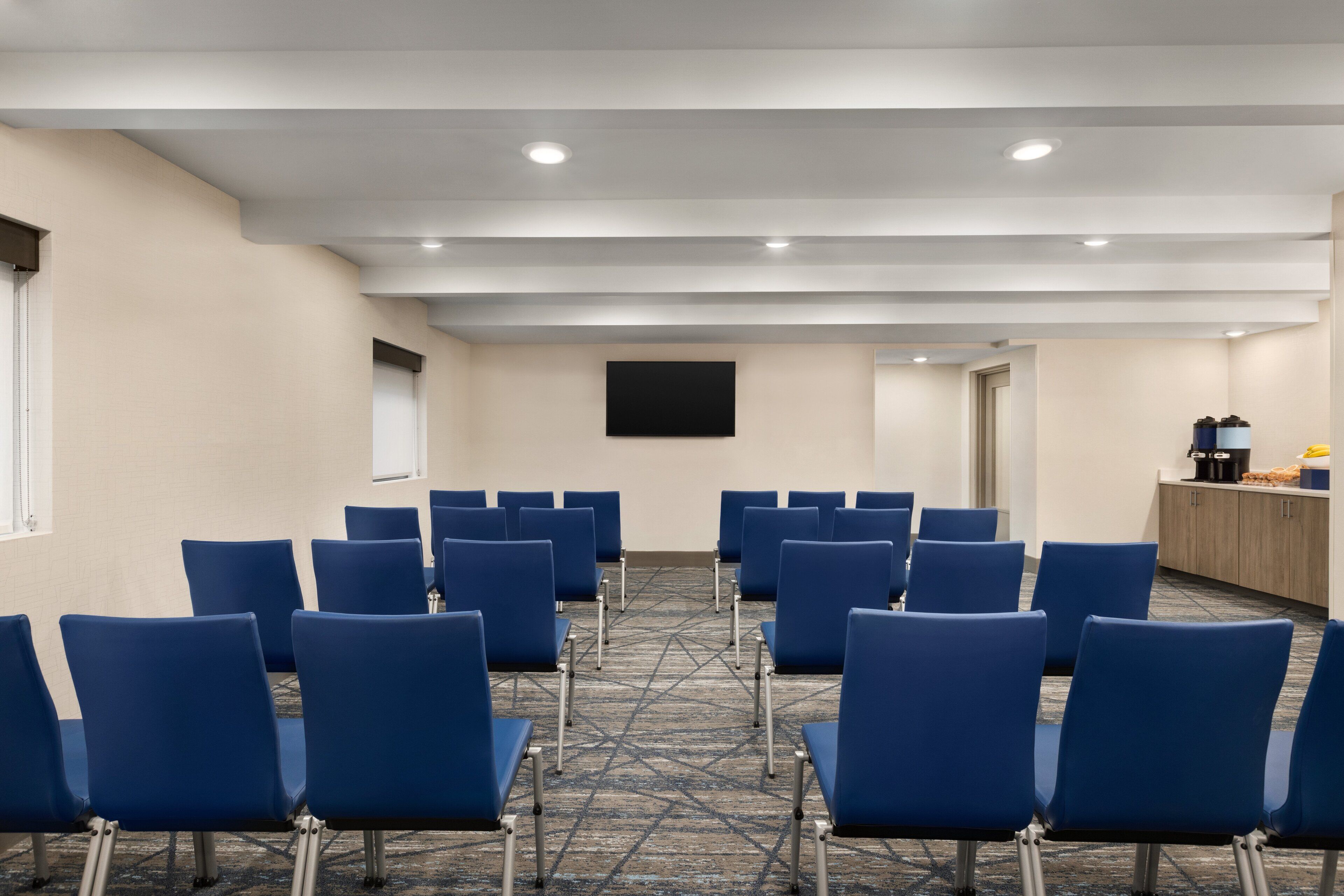 A conference room with rows of blue chairs, a flat-screen TV mounted on the wall, and a kitchenette area with cabinets and appliances.