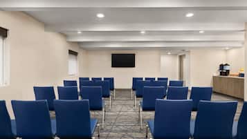 A conference room with rows of blue chairs, a flat-screen TV mounted on the wall, and a kitchenette area with cabinets and appliances.