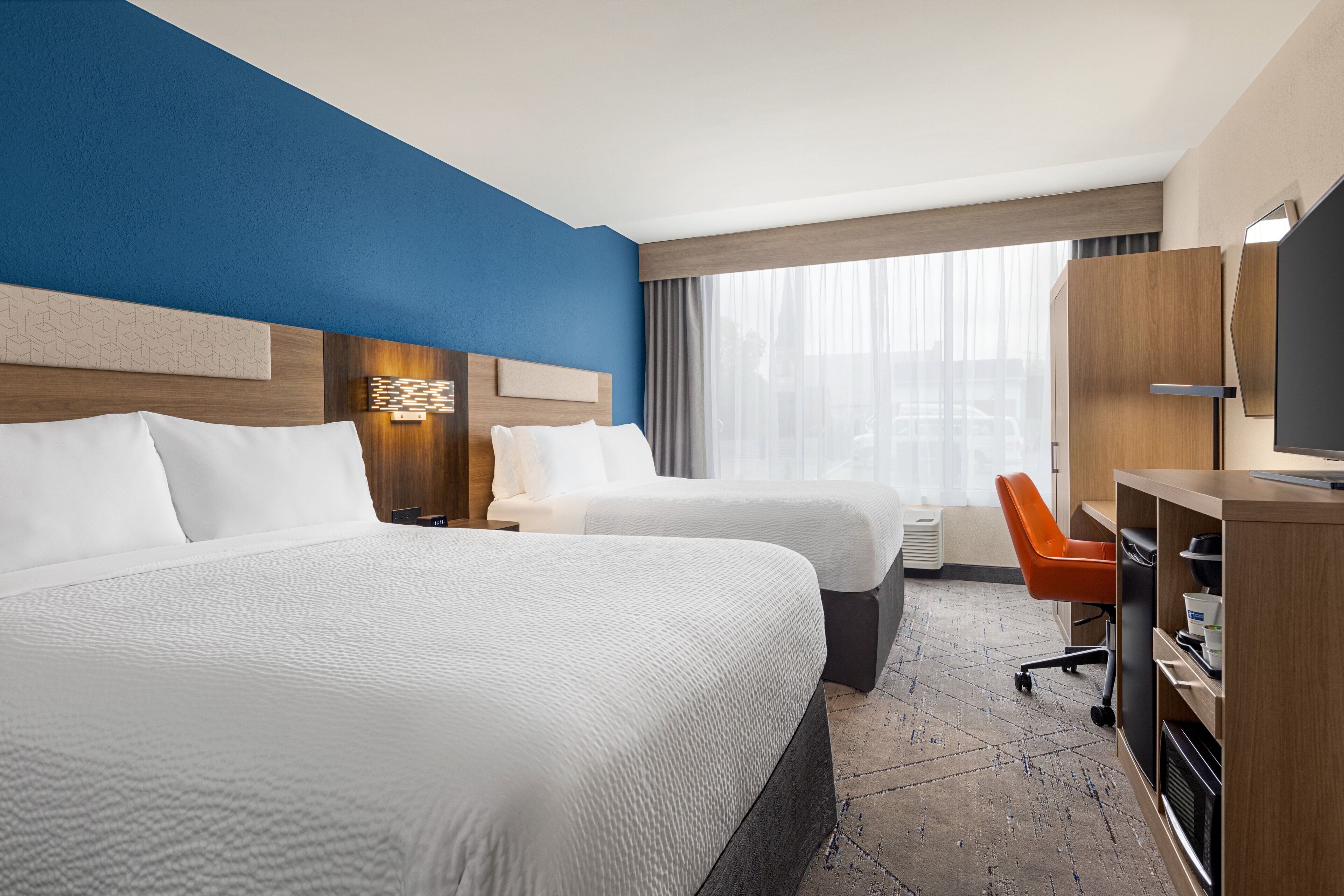 Habitación de hotel con dos camas, un escritorio, una silla naranja y un ventanal con cortinas.