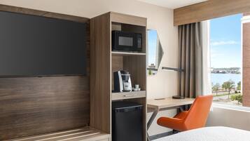 A hotel room with a flat-screen TV, a coffee maker, a microwave, a desk with a chair, and a view of a body of water through the window.