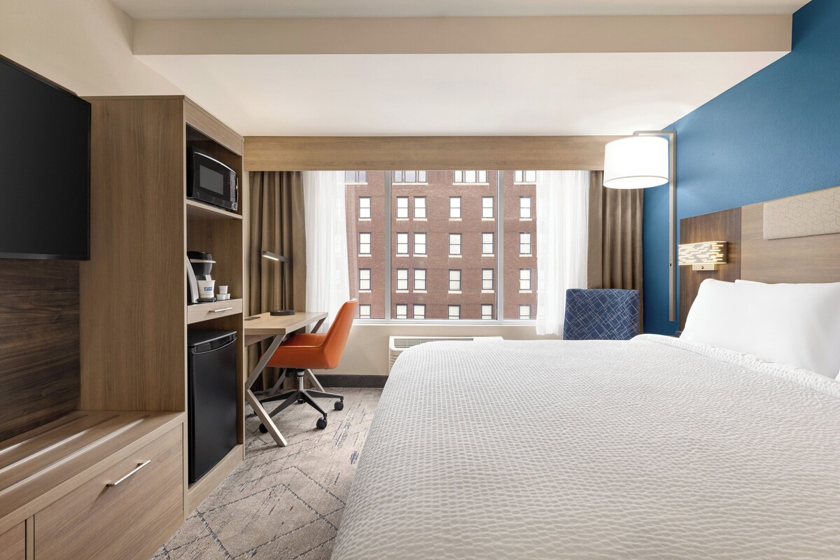 Une chambre d’hôtel avec un grand lit, un bureau avec une chaise, une télévision, un four à micro-ondes et une fenêtre donnant sur la ville.