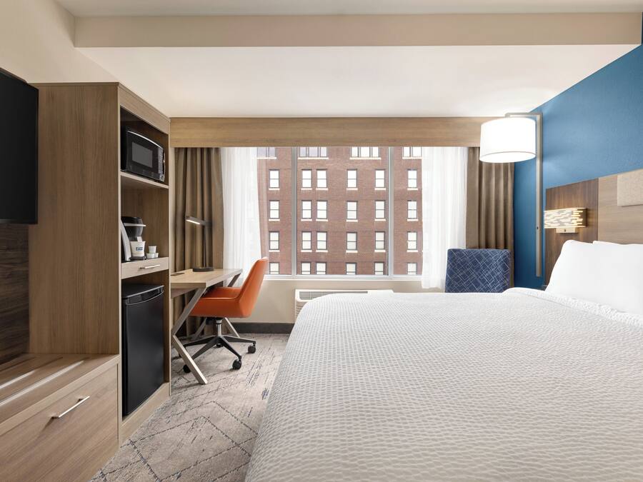A hotel room with a bed, a desk, a chair, a television, a microwave, and a window with a view of a building.