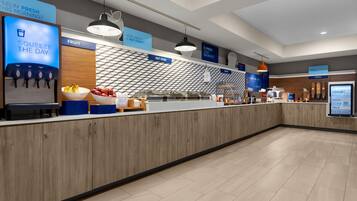 A modern breakfast area with a juice dispenser, fruit display, and labeled sections for hot food, cereal, and yogurt.