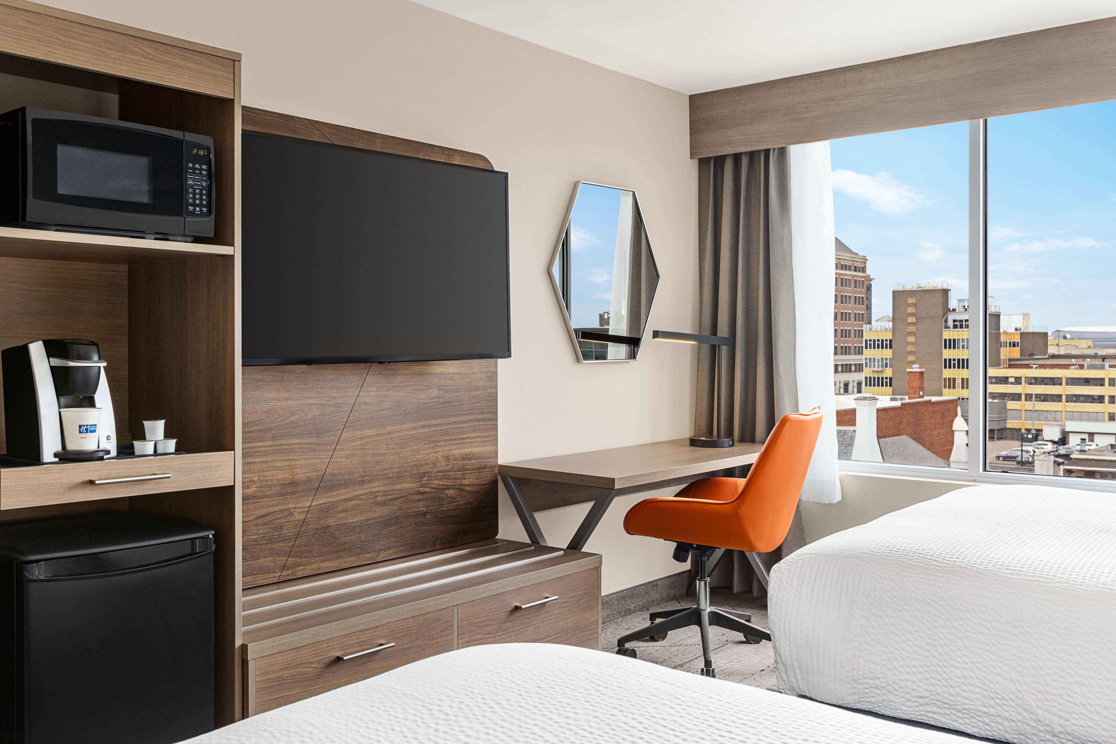 A hotel room with a bed, a desk with a chair, a TV, a coffee machine, and a view of the city.