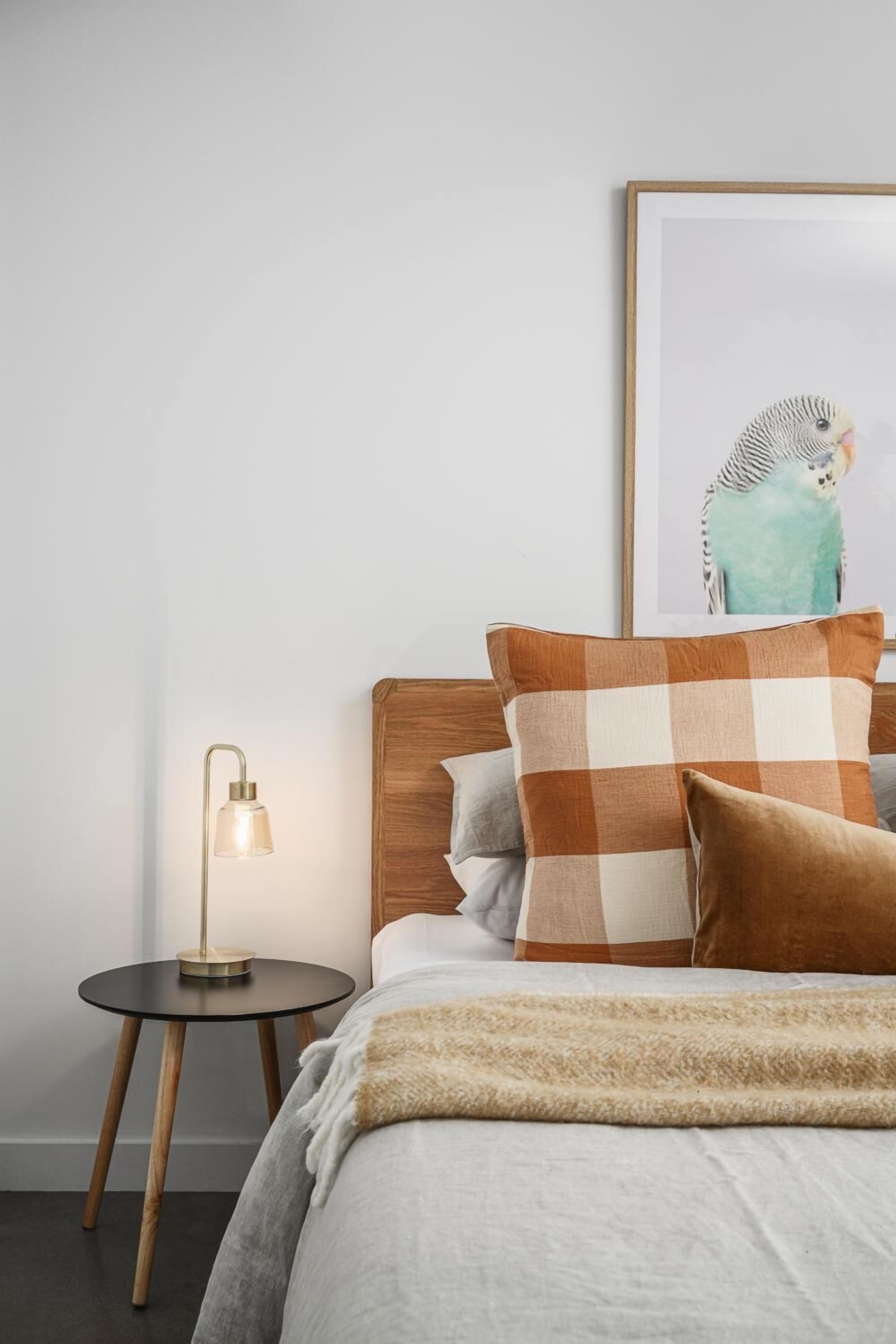 A neatly made bed with a checkered pillow, a small round table with a lamp, and a framed picture of a bird on the wall.
