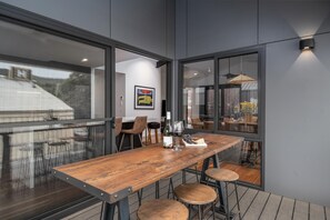 Un espace repas moderne avec une table en bois, des tabourets de bar et une vue sur l’extérieur grâce à de grandes fenêtres.