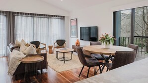 Una sala de estar moderna con un sofá, sillas, una mesa de centro y una mesa de comedor. Hay un televisor montado en la pared y un ventanal con cortinas.