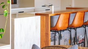 A modern kitchen with a marble countertop, wooden bar counter, and bar stools. There are pendant lights above the counter and a plant on the left. - 25 Walkers Lane - Luxurious living with breath taking views (Bright)