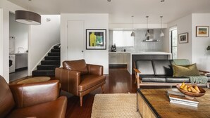 A modern living room with leather sofas, a coffee table with books and fruit, and a staircase leading to a kitchen with a view.