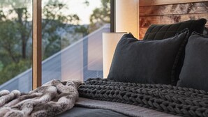 A modern bedroom with a large window, a bed with dark bedding, and a bedside lamp. - The Grove Estate - The pièce de resistance of this property is the view, be it the 360-degree views of the surrounding bush side, valley and the Region’s famous Mt Buffalo or perhaps the view of the spectacular and famous artwork showcased through th (Porepunkah)