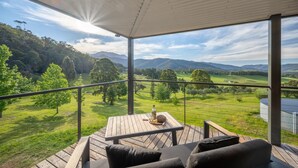 A balcony with a wooden table and chairs, overlooking a green landscape with trees and hills. - The Grove Estate - The pièce de resistance of this property is the view, be it the 360-degree views of the surrounding bush side, valley and the Region’s famous Mt Buffalo or perhaps the view of the spectacular and famous artwork showcased through th (Porepunkah)
