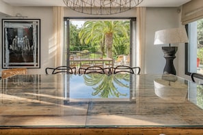 A modern dining area with a glass table, chairs, a lamp, and a view of a tropical garden.