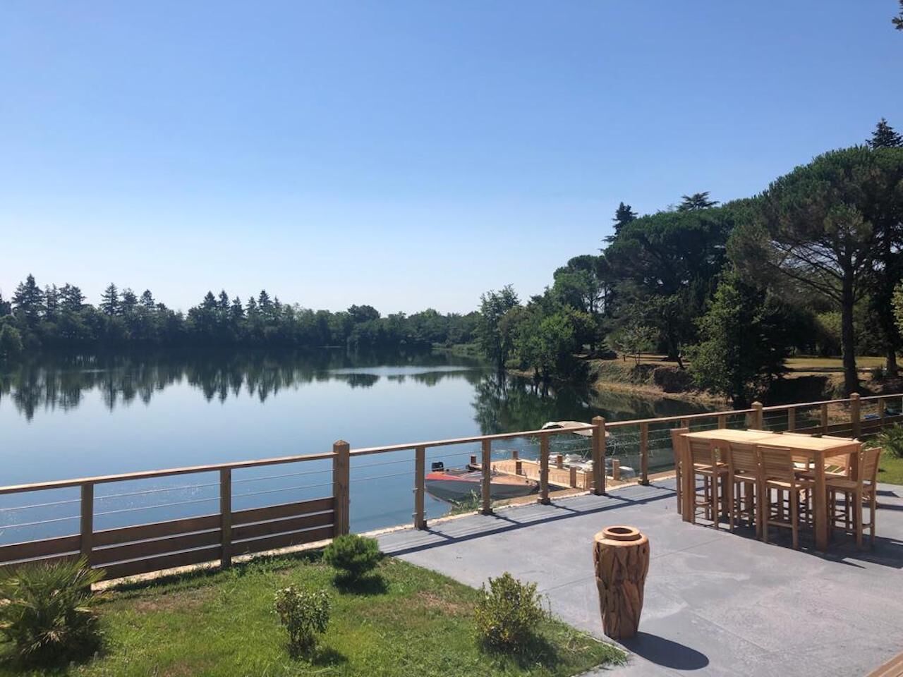 En patio ved søen med træterrasse, et bord og stole, samt udsigt over søen og de omkringliggende træer.