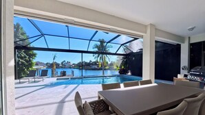 Ein Poolbereich mit Tisch und Stühlen, einem Grill und Blick auf einen See mit Palmen.