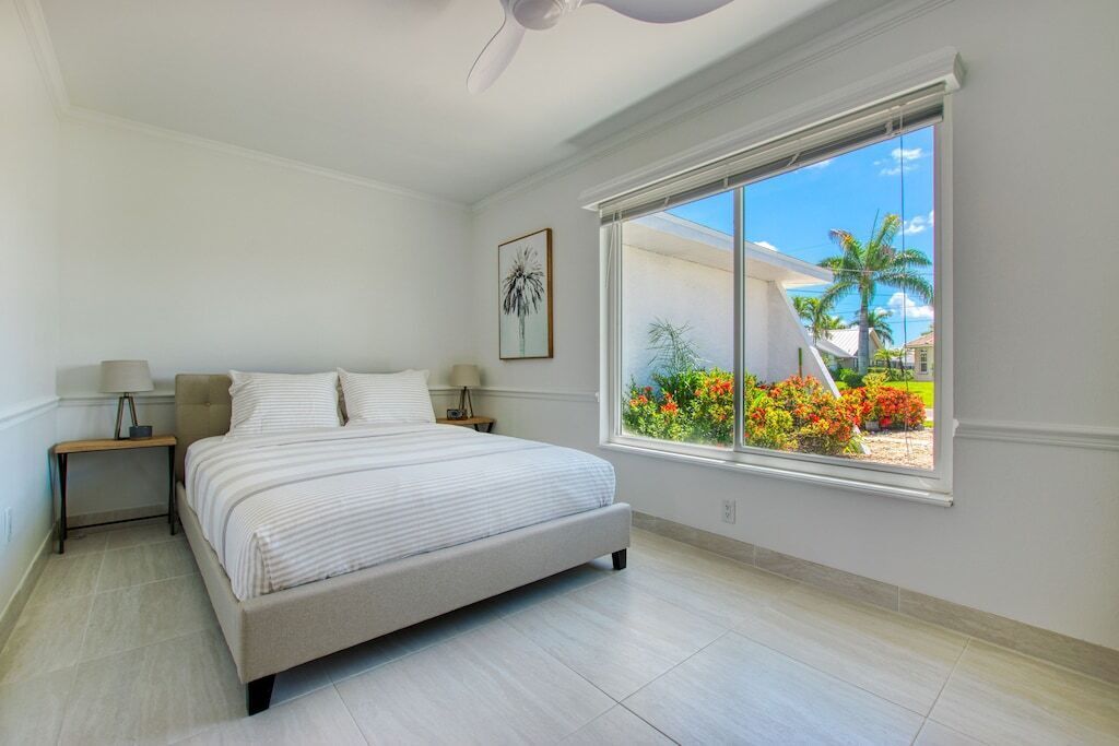 A bedroom with a large bed, a nightstand, a lamp, a ceiling fan, and a window with a view of a garden and buildings.