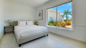 Ein Schlafzimmer mit einem großen Bett, einem Nachttisch, einer Lampe, einem Deckenventilator und einem Fenster mit Blick auf einen Garten und Gebäude.