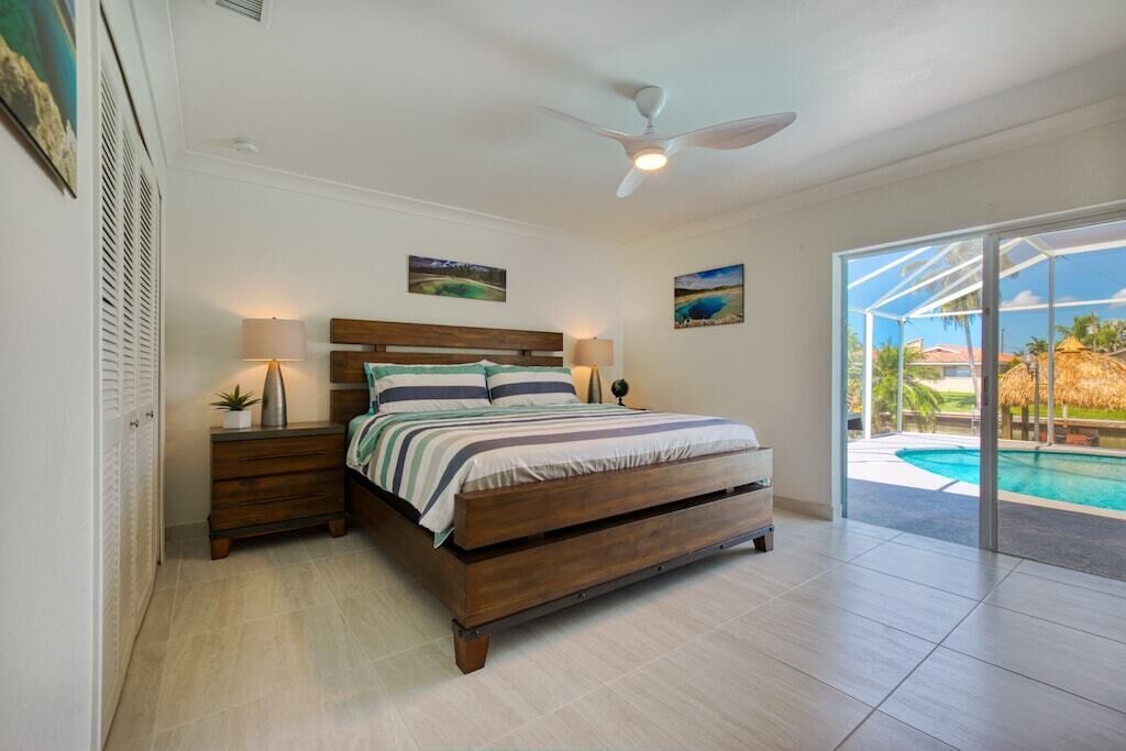 A bedroom with a wooden bed, two bedside tables, a ceiling fan, and a view of a swimming pool.