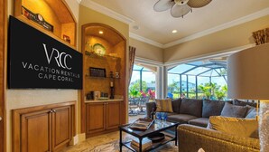 Un salón amplio con un televisor grande mostrando 'VACATION RENTALS CAPE CORAL', una zona de estar acogedora y vistas al exterior a través de un ventanal.