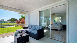 A covered outdoor area with a sofa and a table, a sliding glass door leading to a bedroom, and a view of a green landscape.
