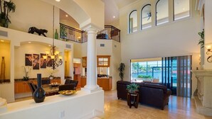 A spacious living area with a high ceiling, a balcony, and a kitchen in the background.