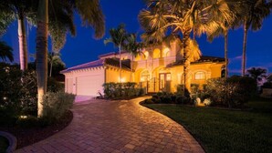 A well-lit, two-story house with a driveway leading to a garage, surrounded by palm trees and a landscaped garden.