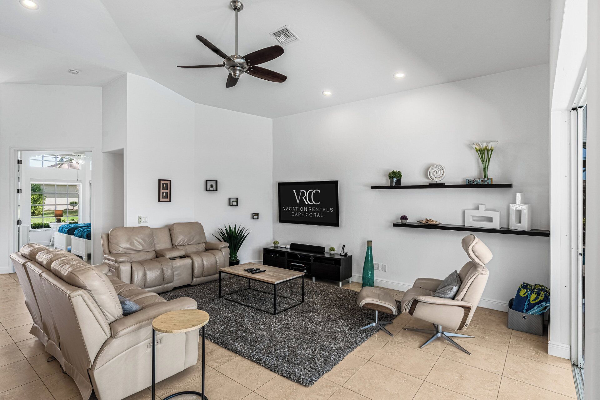Un salon moderne avec un canapé en cuir beige, une table basse, un téléviseur et un ventilateur de plafond. On y trouve des étagères ornées d’objets décoratifs et une porte qui donne sur une autre pièce.