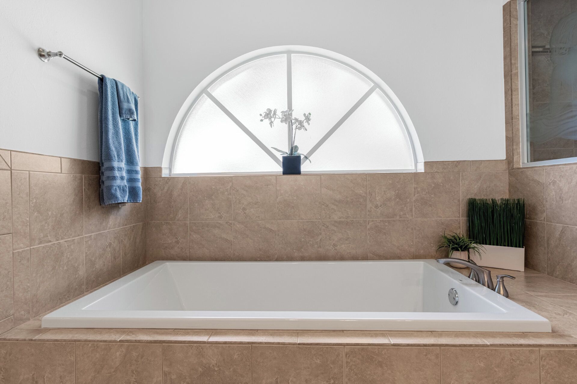 A modern bathroom with a freestanding bathtub, a towel rack with a blue towel, a potted plant, and a window with a decorative glass design.