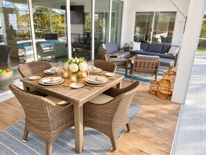 An outdoor dining area with wicker chairs, a wooden table set for four, and a view of a pool and lounge area in the background.