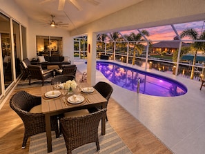 Un espacio exterior para comer con piscina, mobiliario de mimbre y vista a una puesta de sol entre palmeras.