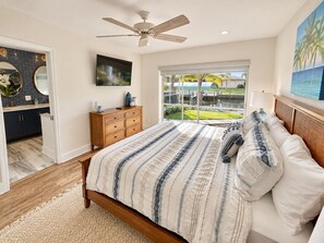 A bedroom with a bed, a ceiling fan, a TV, a dresser, and a view of a water body and greenery.