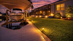 Ein Boot an einem Steg, ein Haus mit einer Veranda und eine gut beleuchtete Rasenfläche in der Abenddämmerung.