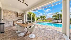 A modern kitchen with a stone backsplash, a dining area with a wooden table and white chairs, and a poolside view with a clear sky.