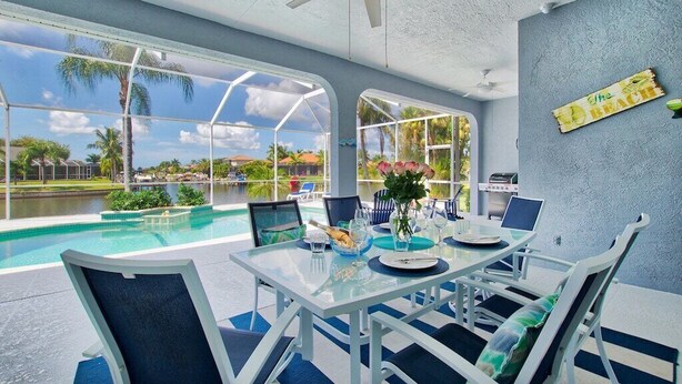 A dining area with a glass table, white chairs, and a view of a swimming pool and palm trees. - VRCC Villa Sansibar (Cape Coral)
