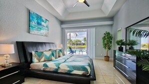 A bedroom with a bed, a ceiling fan, a nightstand, a dresser, and a large window. - VRCC Villa Sansibar (Cape Coral)