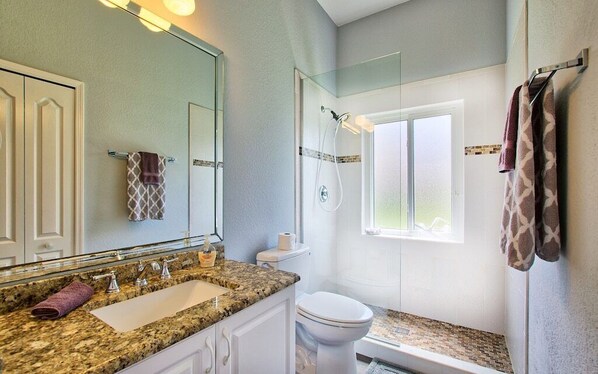 Een badkamer met een wastafel op een granieten blad, een wit toilet, een douchecabine van glas en een raam.