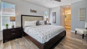 Ein Schlafzimmer mit Bett, Nachttischen, Deckenventilator, Fenster mit Jalousien, Wandbild und einem durchgehenden Türrahmen sichtbaren Badezimmer.