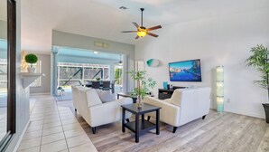 Ein modernes Wohnzimmer mit einer Couch, einem Couchtisch und einem Fernseher. Es gibt einen Deckenventilator, ein großes Fenster und einen Blick ins Freie.