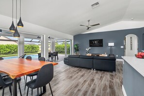 Una sala de estar moderna con un sofá gris, una mesa de comedor de madera y sillas negras. Hay un televisor montado en la pared y un ventilador de techo.