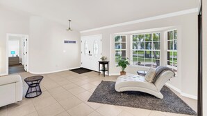Ein geräumiger Wohnbereich mit einem weißen Sofa, einem runden Tisch und einem großen Fenster mit Jalousien.