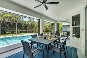 Area pranzo a bordo piscina con tavolo apparecchiato per quattro, ventilatore a soffitto e vista sulla piscina e sul verde.