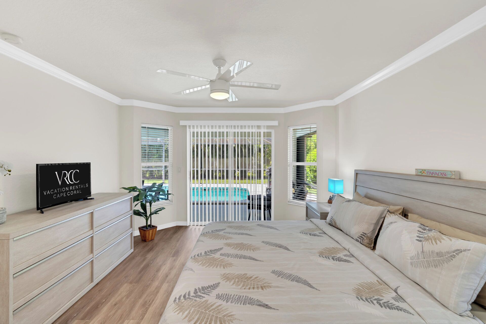 A bedroom with a bed, a ceiling fan, a TV, a nightstand, and a view of a swimming pool.