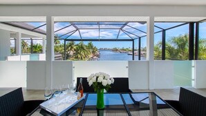 Ein stilvoller Essbereich mit einem Glastisch, weißen Stühlen und einer Vase mit weißen Blumen. Auf dem Tisch steht eine Weinflasche und Gläser. Der Raum verfügt über große Fenster mit Blick auf ein Gewässer und Palmen.