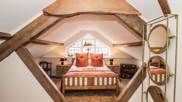 Un dormitorio con techo inclinado, cama con ropa de cama blanca, cabecera de madera, ventana con vitrales y un espejo en la pared.